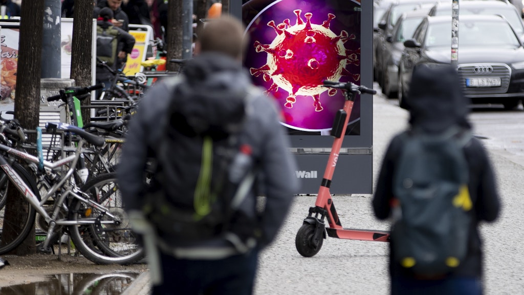 Passanten gehen vor einer elektronischen Werbetafel, auf der eine Illustration des Coronavirus zu sehen ist. (Symbolbild)