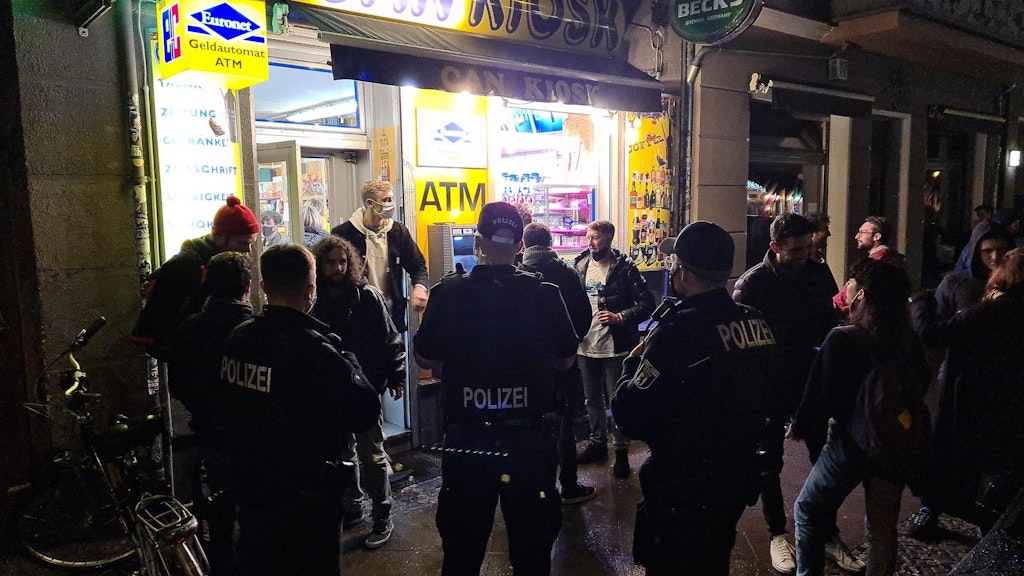 Drei Polizisten stehen vor einem Späti in Friedrichshain, der trotz Sperrstunde geöffnet hat und gut besucht ist.