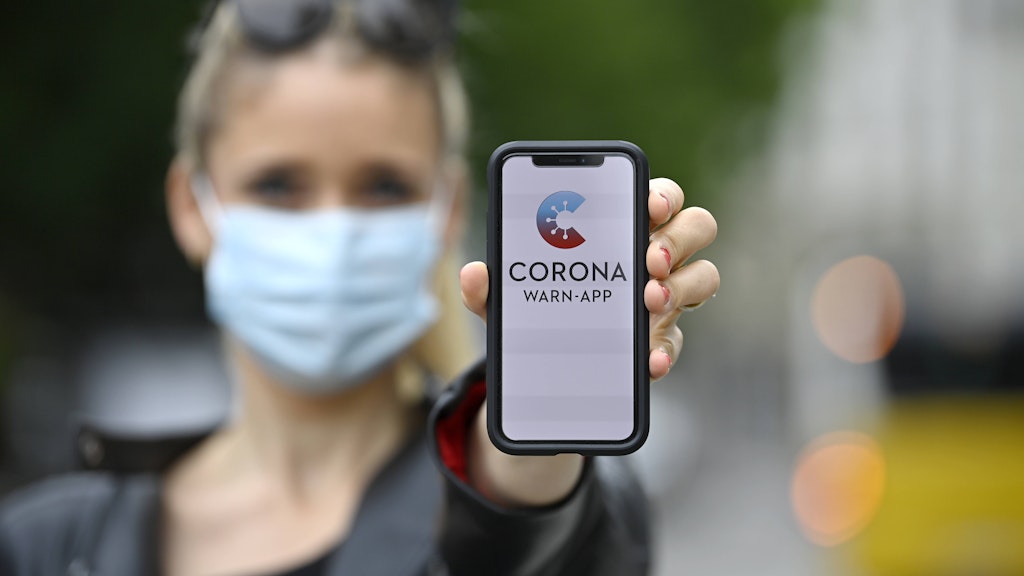 Frau mit Mundschutzmaske, zeigt Smartphone mit Corona Warn-App.