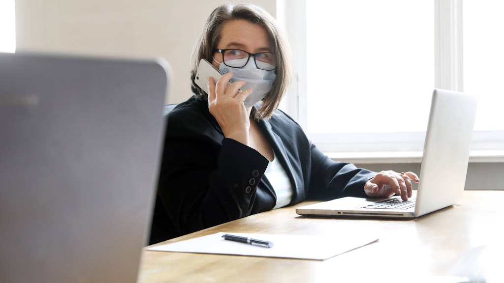 Frau im Büro am Arbeitsplatz mit Mundschutz, um Ansteckung bei Kollegen zu vermeiden.
