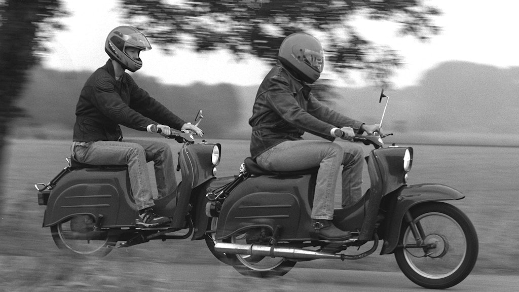 Zwei junge Männer fahren mit ihren Schwalbe-Rollern über einen Feldweg. Die Schwalbe ist nicht irgendein Moped, sondern ein vielverkauftes Zweirad aus dem Suhler Volkseigenen Betrieb (VEB) Fahrzeug- und Jagdwaffenwerk Simson. <br>